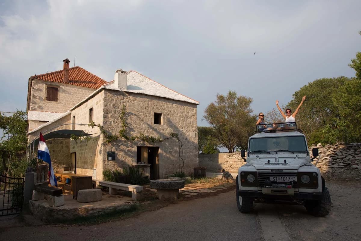 Two visitors on the roof of a white jeep beside a stone house.