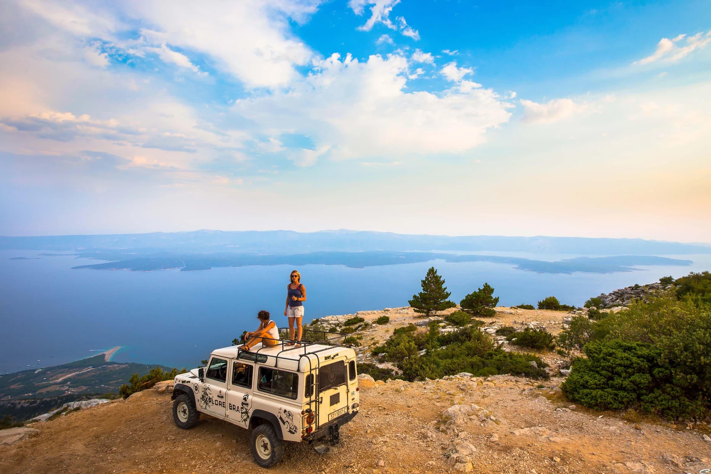Two visitors on the roof of a white jeep at a rocky viewpoint above the Adriatic.