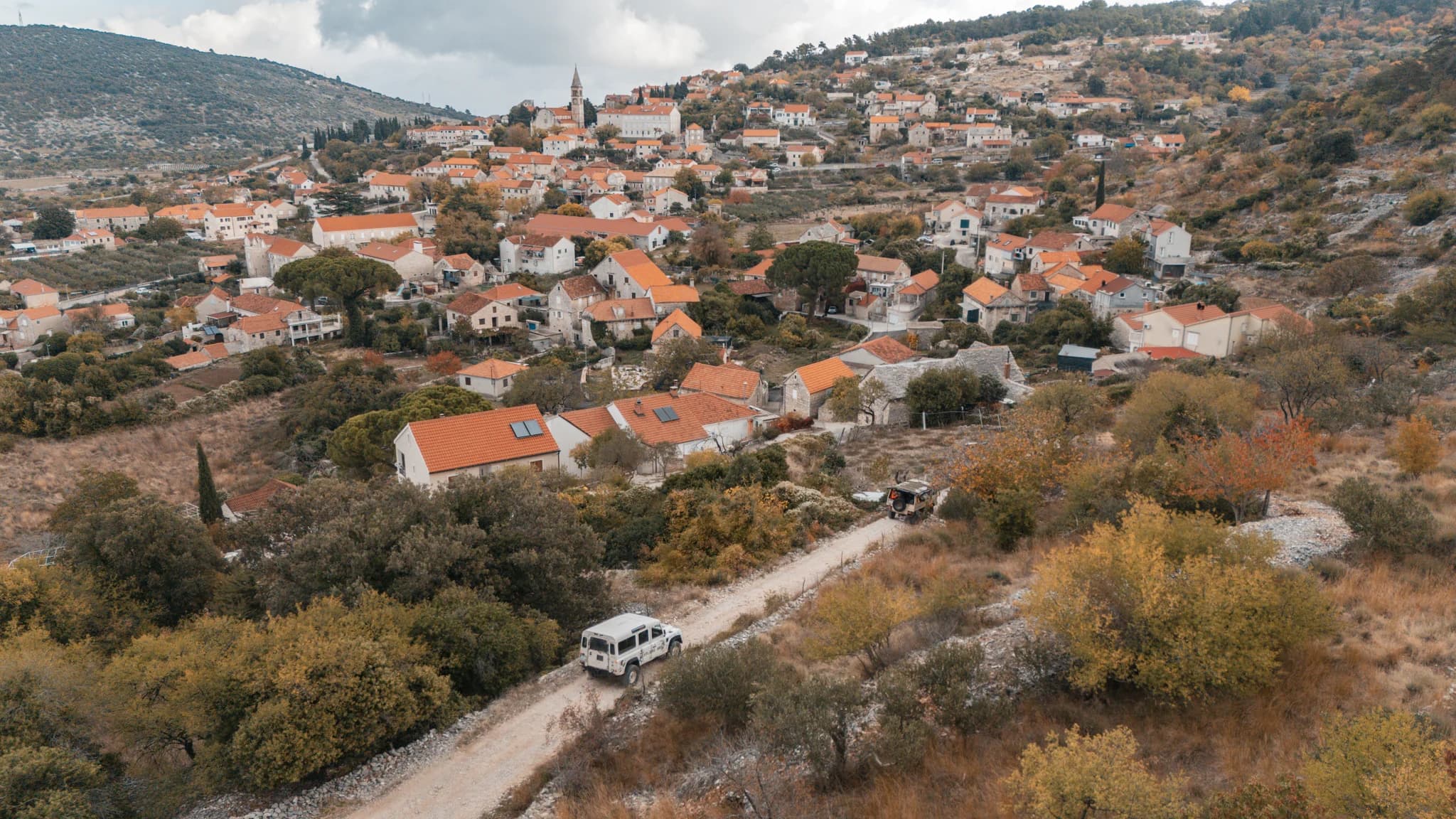 A white jeep on a road below a hillside village with stone houses and red roofs.