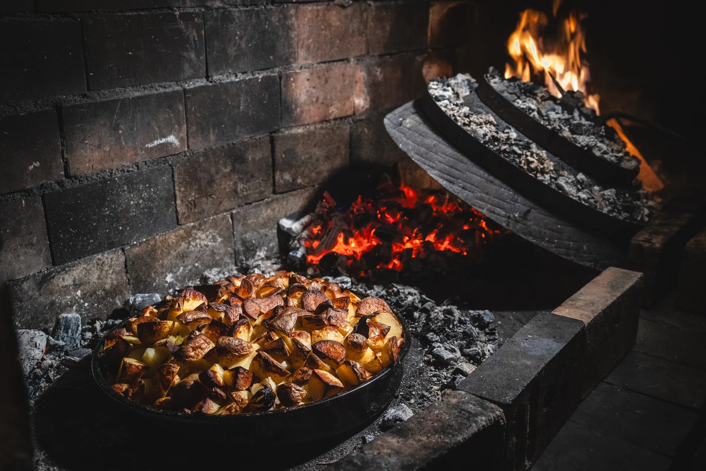 A pan of peka cooking over embers beside its heavy lid in a stone hearth.