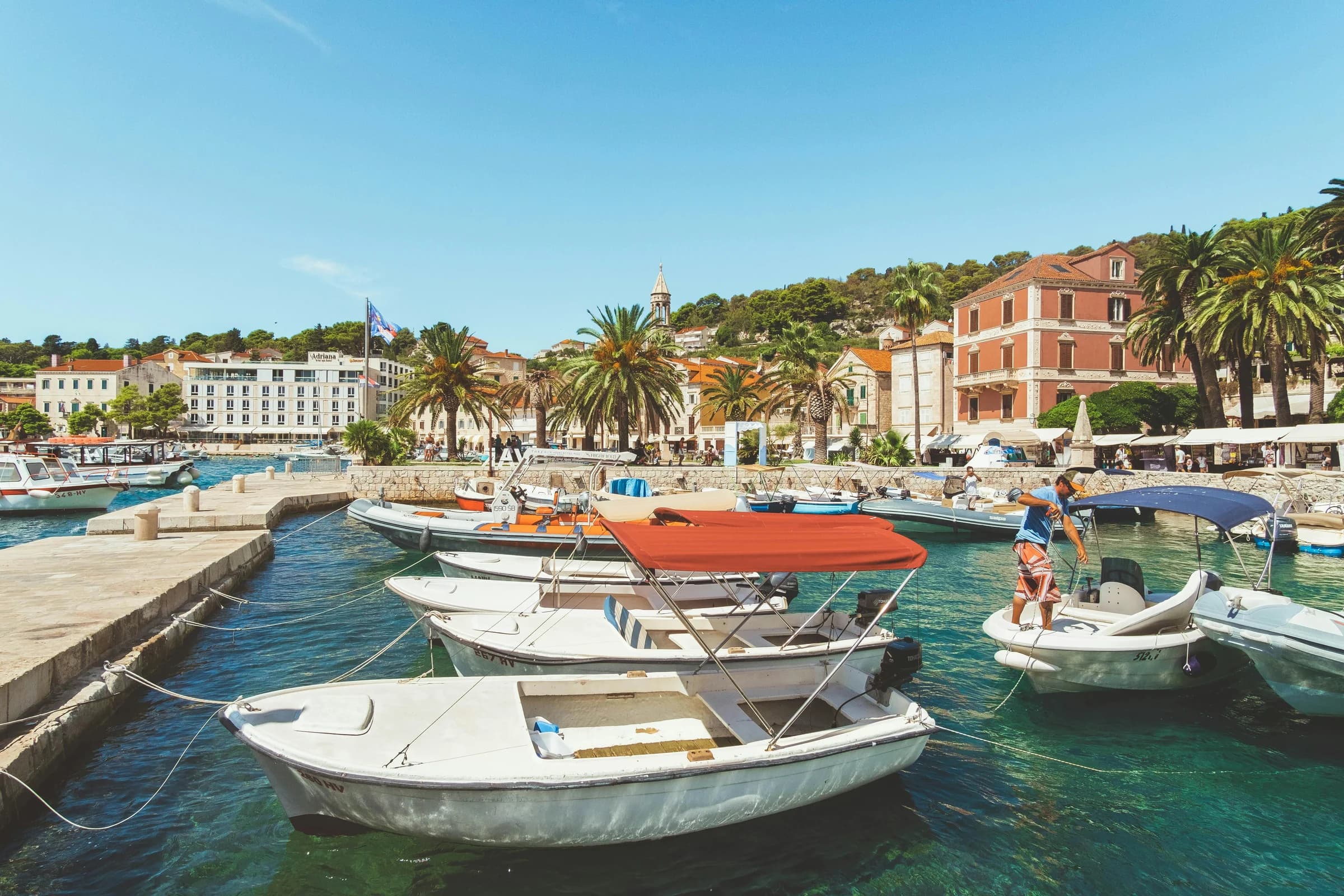 Small boats moored along Hvar's stone waterfront beneath palm trees and red-roofed buildings.