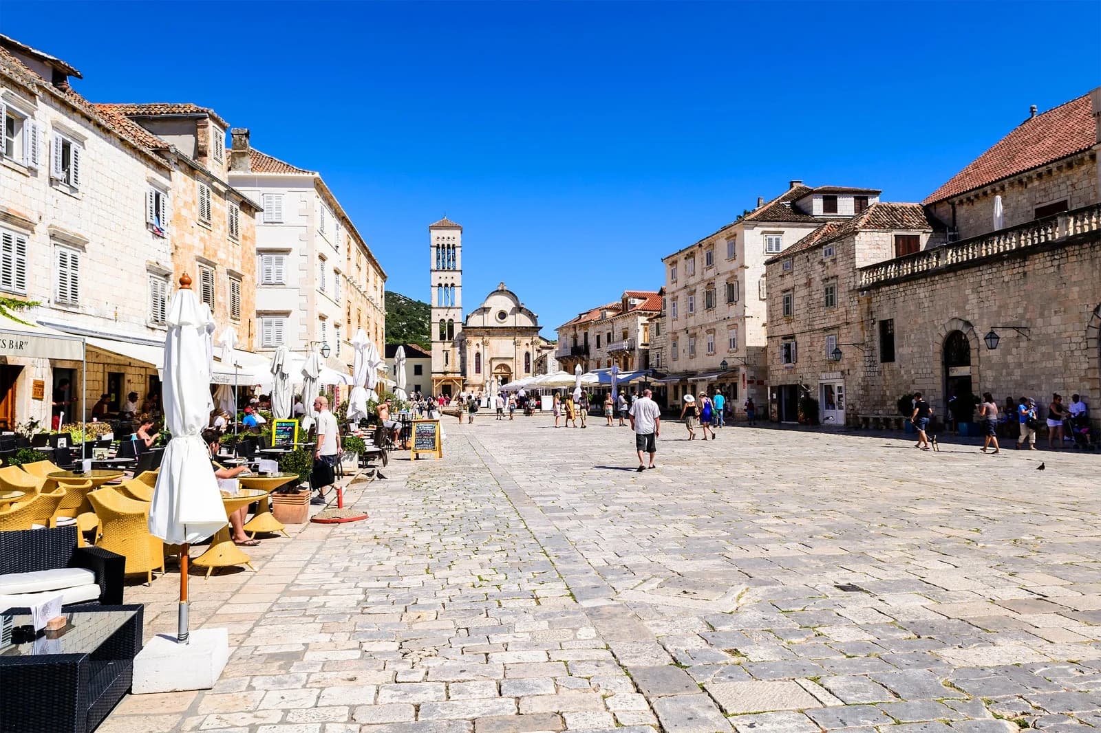People walking through Hvar’s stone-paved square between old buildings and a church tower.