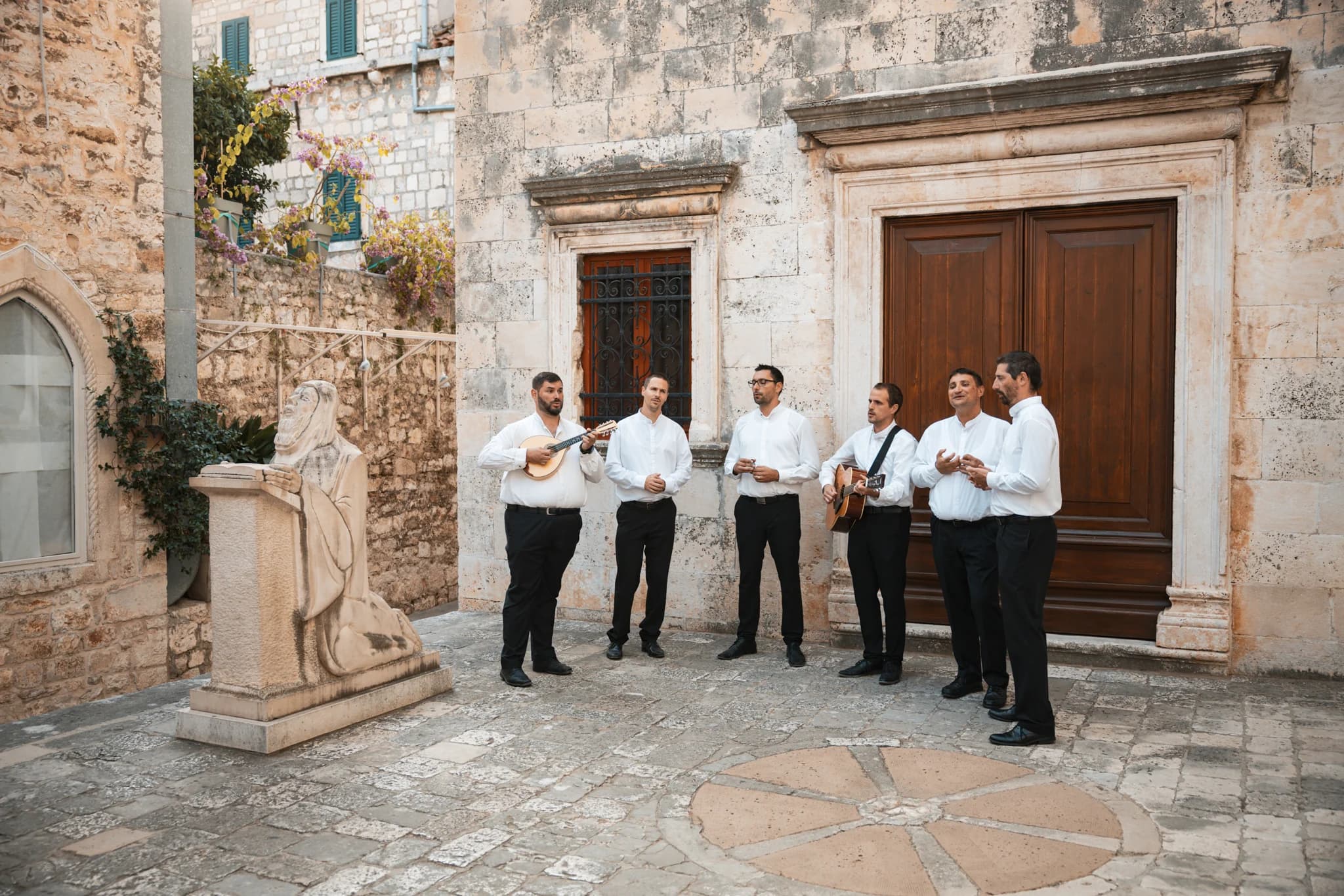 Six men singing and playing stringed instruments outside a stone building.