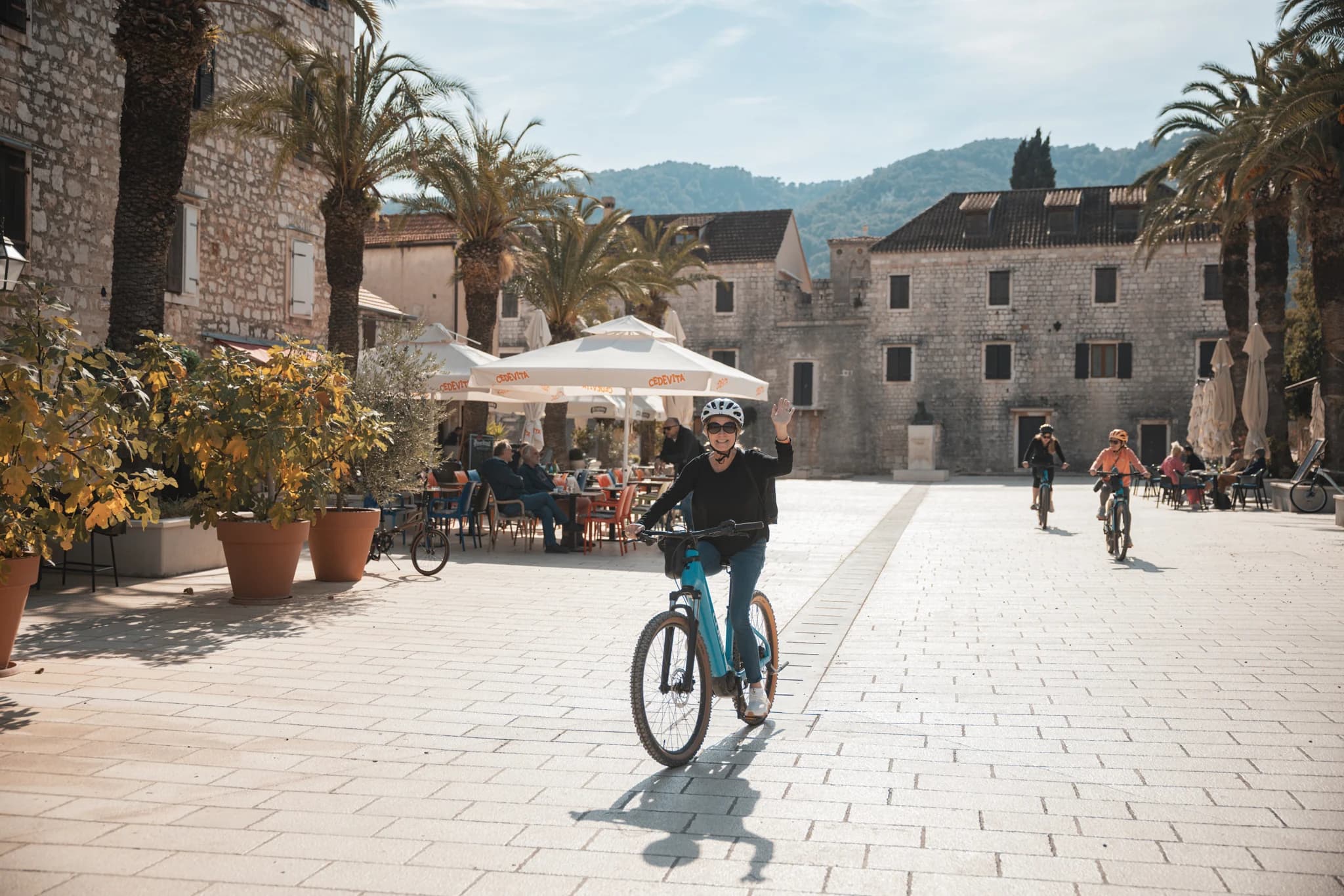 Helmeted cyclists with bicycles in a stone-paved square lined by old buildings.