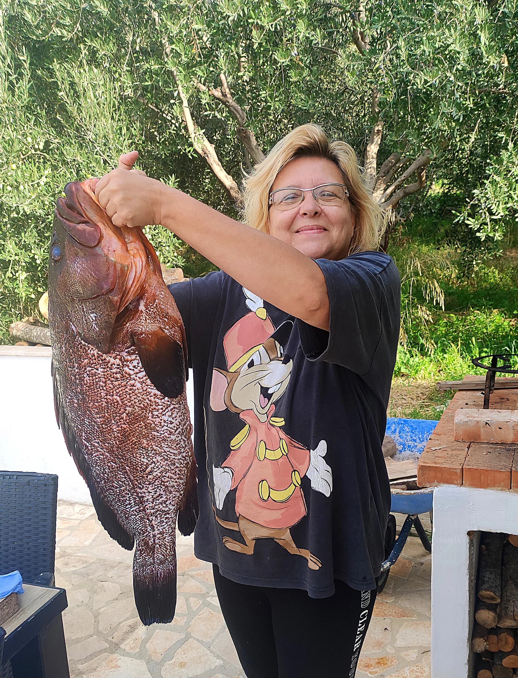 A woman holding a large fish beside olive trees and an outdoor cooking area.