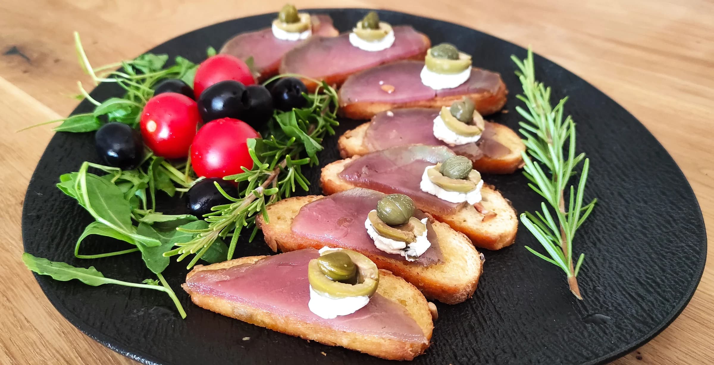 Slices of bread with cured meat, olives and garnish arranged on a dark plate.