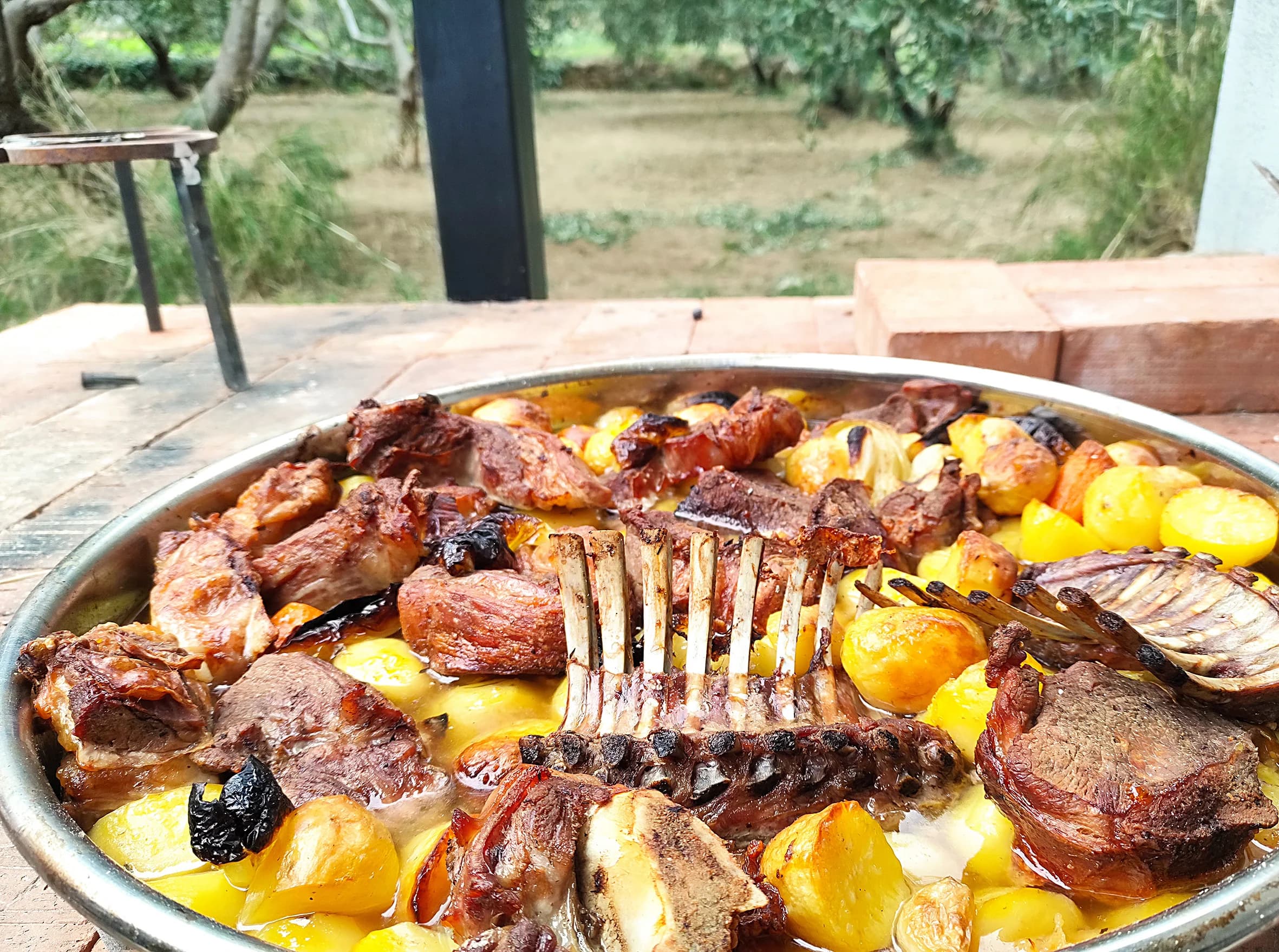 A large platter of roasted food and potatoes on a table beside olive trees.