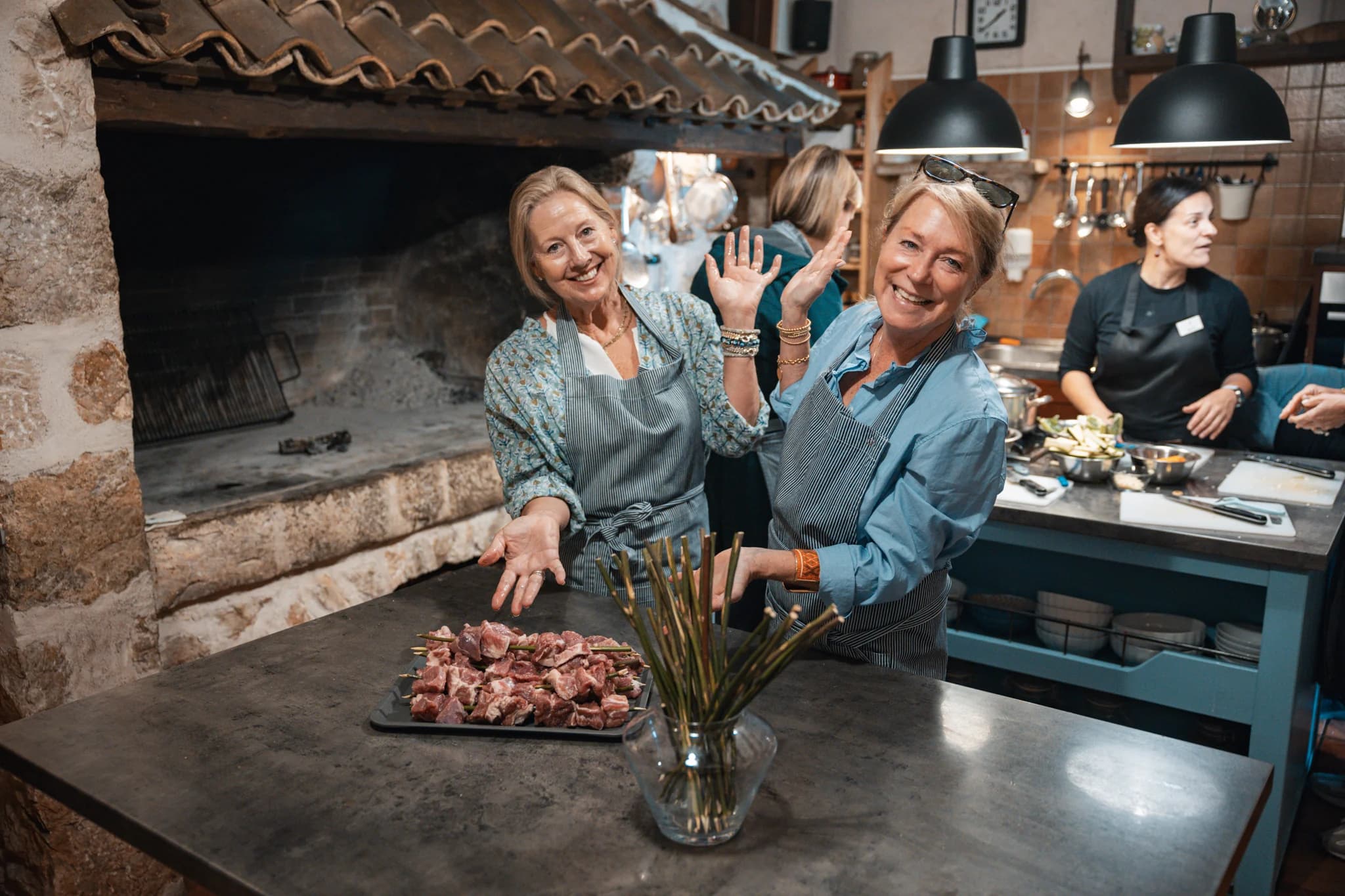 Two cooking-class participants wearing aprons beside a stone hearth and a tray of skewers.