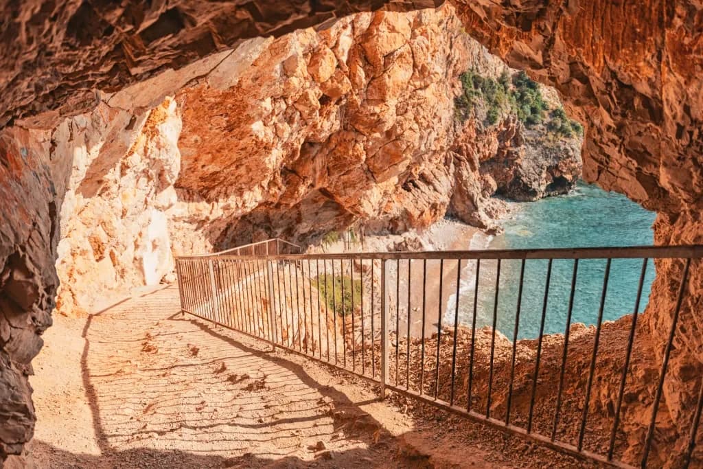 A railed stone path beneath an orange rock overhang beside the sea.