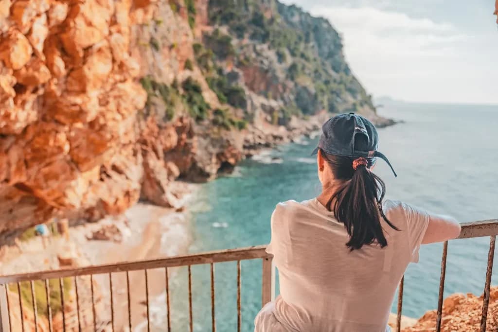 A visitor in a cap looking out across the sea from a railed path beneath coastal cliffs.