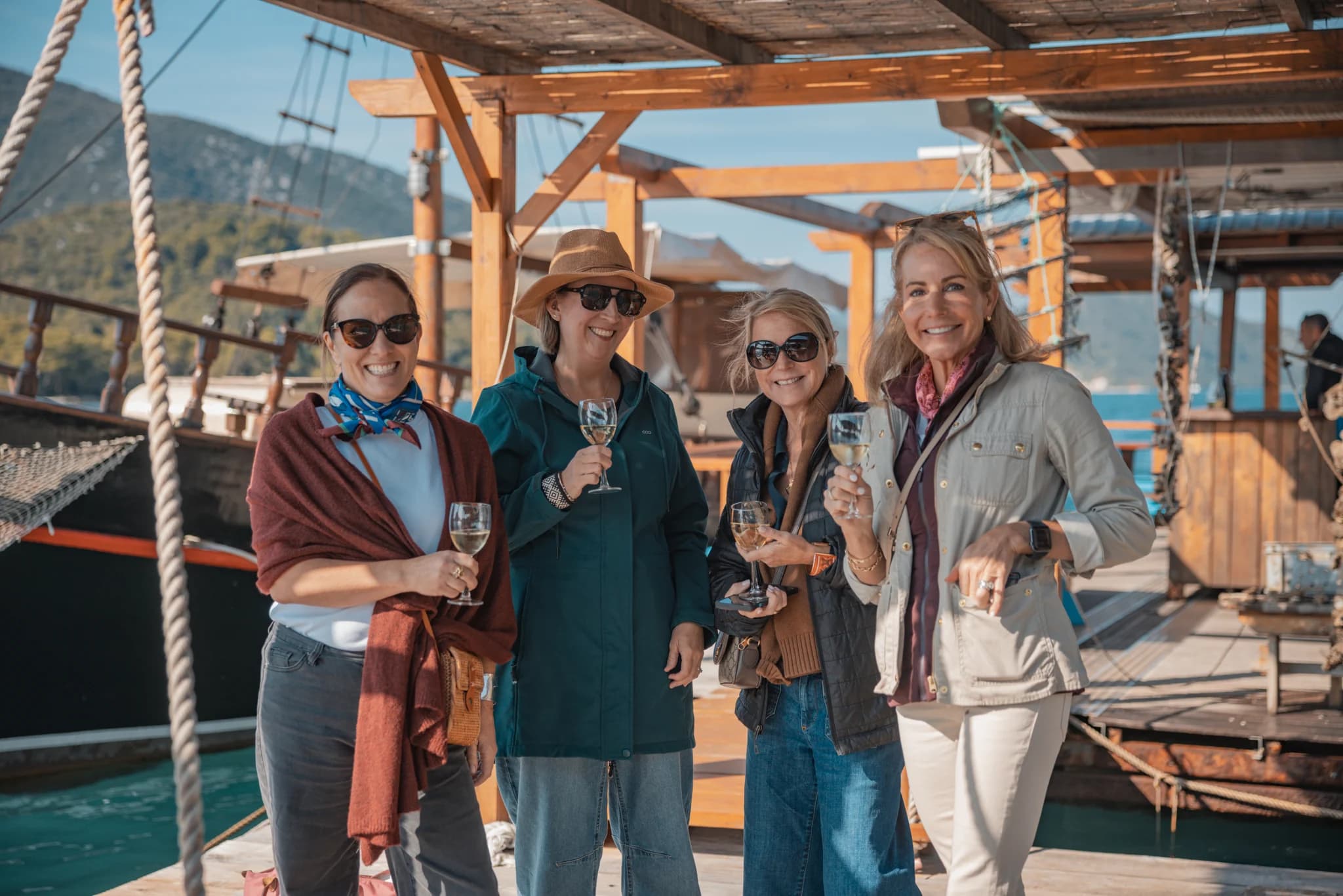 Four people holding glasses on a wooden deck beside the water.