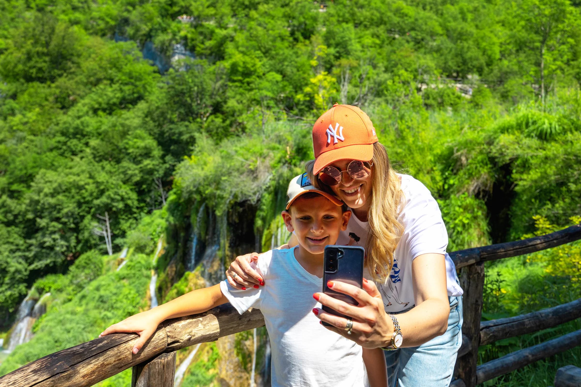 Two visitors taking a photograph beside a wooden railing, with forest and waterfalls behind them.