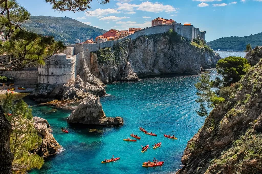 Kayaks crossing a blue inlet beneath stone fortifications on a high cliff.