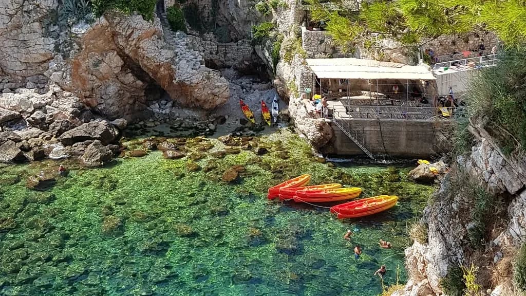 Orange and yellow kayaks in clear water beside a rocky shoreline.