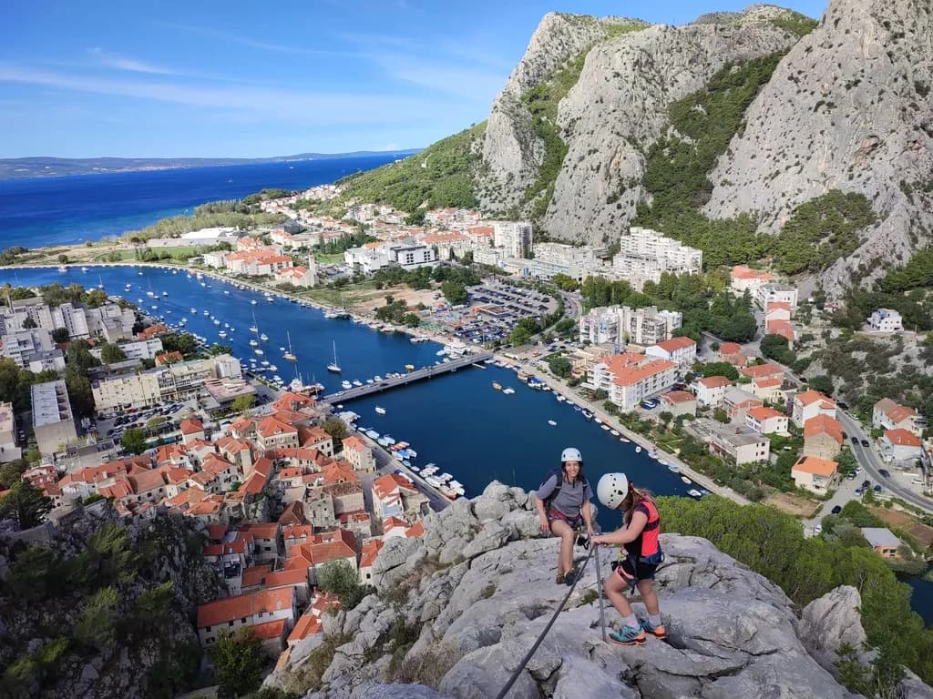 Two helmeted climbers on a rocky ledge above a river and red-roofed buildings.