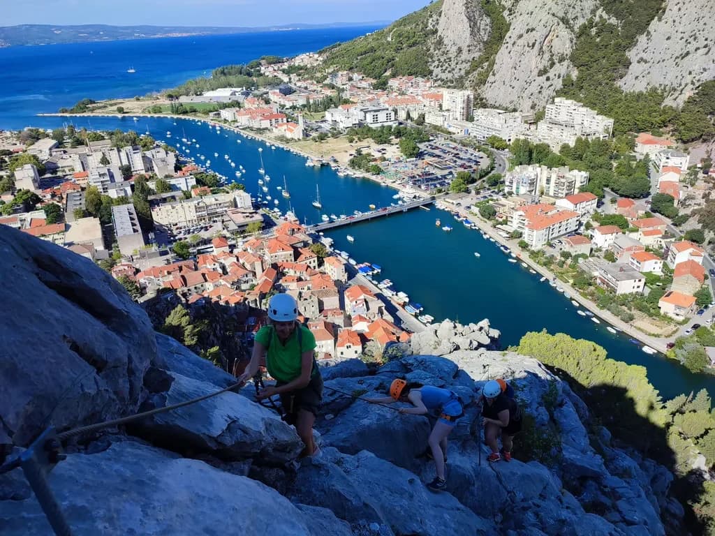 Helmeted climbers following a fixed cable across a rocky face above a river and town.
