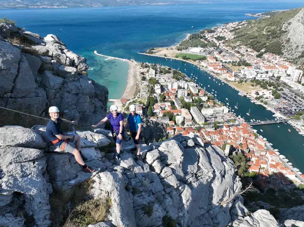 Three helmeted climbers on a rocky ledge above a town and winding river.