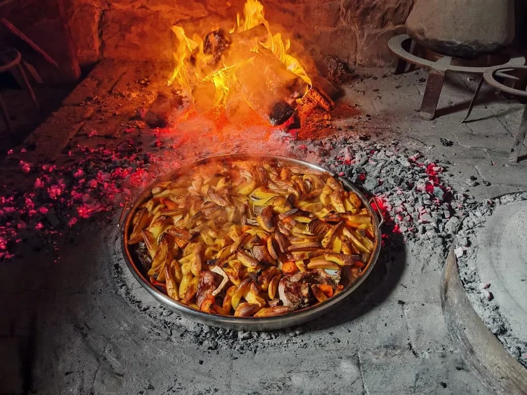 A pan of roasted potatoes and meat beside a wood fire and glowing embers.