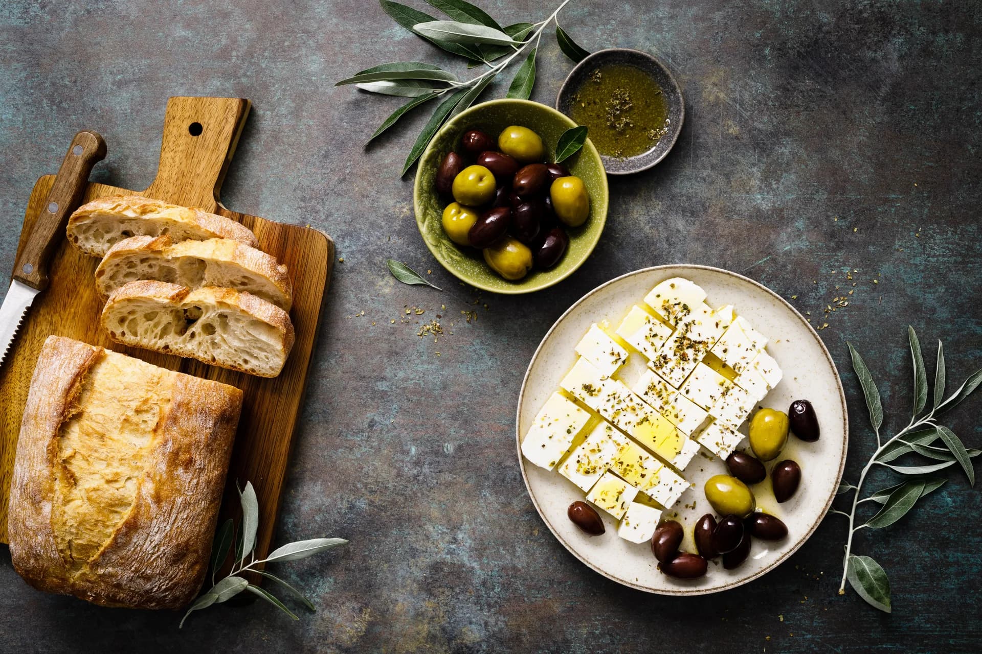Bread, olives, olive oil, and cheese arranged on a stone tabletop.