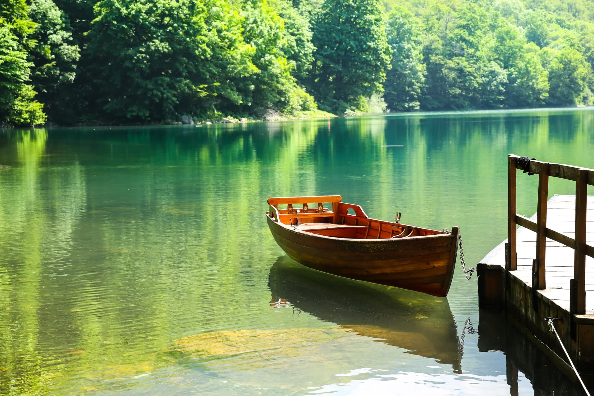 A wooden rowing boat rests beside a jetty on a still, tree-lined lake.